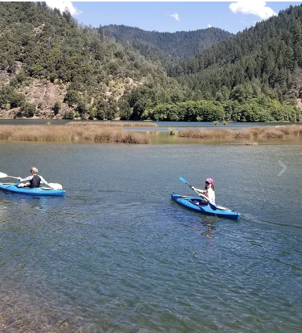 Two kayakers paddling calm water below forested hills